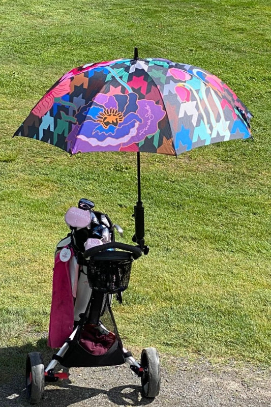A colorful floral golf umbrella with a houndstooth pattern, displayed open on a grassy surface, with a pink golf bag and umbrella stand in the background.