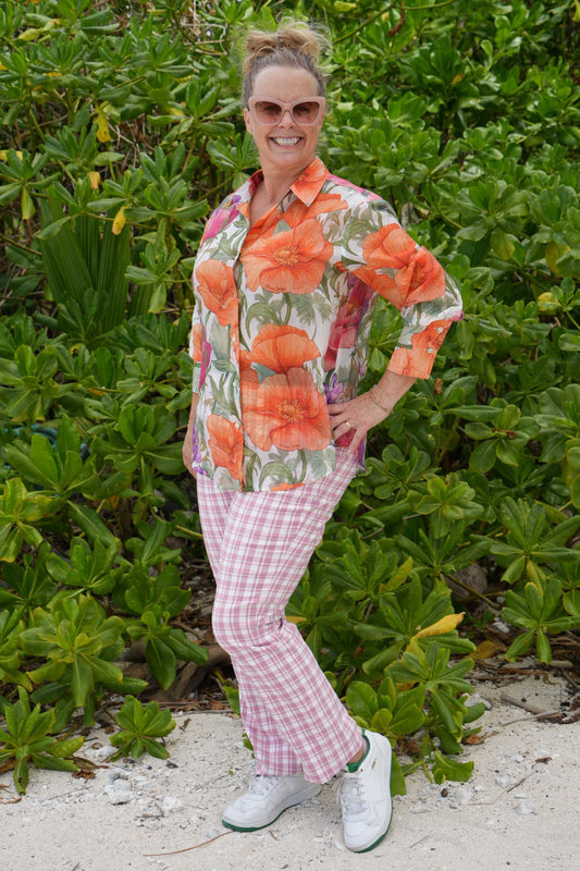 Woman in a colorful floral shirt and pink pants standing in front of green foliage.