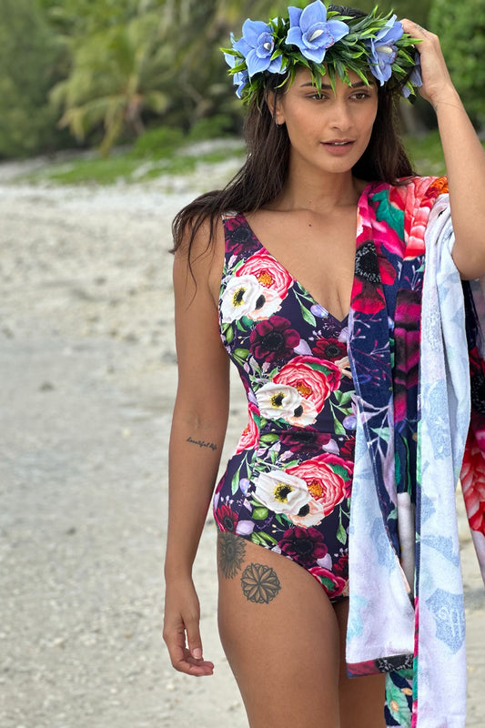 A woman wearing a navy swimsuit with a poppy floral pattern, standing on a beach with a floral crown and towels in the background.