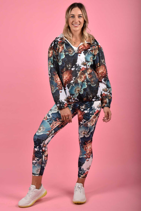 A woman standing against a pink background, wearing blue floral yoga leggings and a matching top with long sleeves.