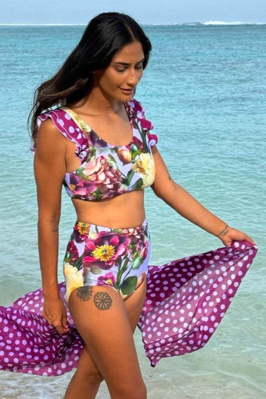 A woman at the beach wearing a two-piece bikini swimsuit with a floral lilac pattern and spot accents.