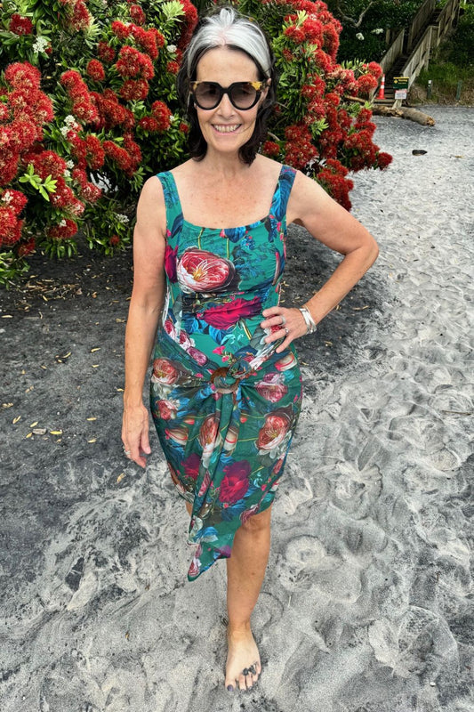 A person wearing a multicolored floral swim scarf wrap with a predominantly green and pink pattern, standing in front of a bush with red flowers.
