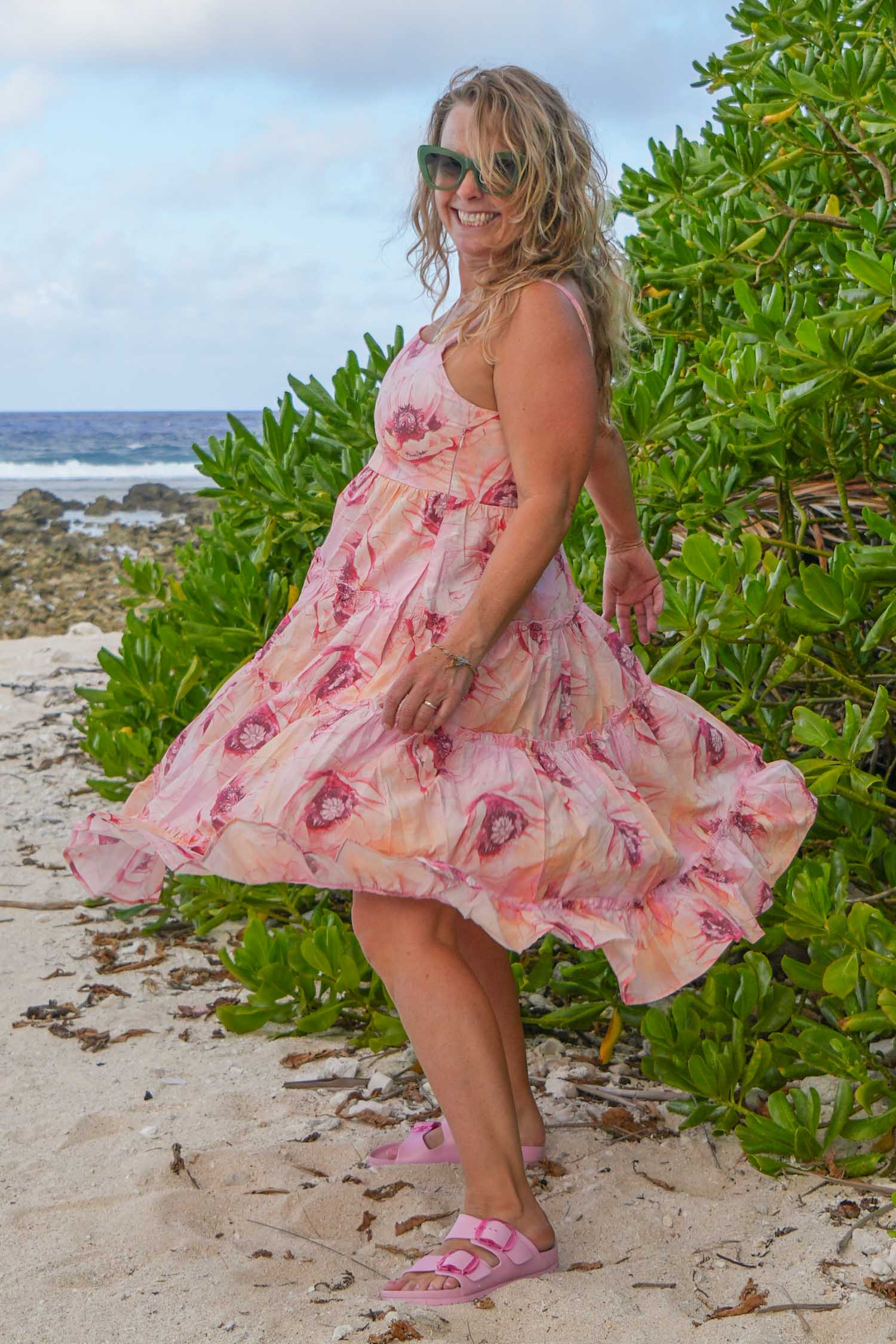 Woman in a pink floral dress standing on a beach with greenery and ocean in the background