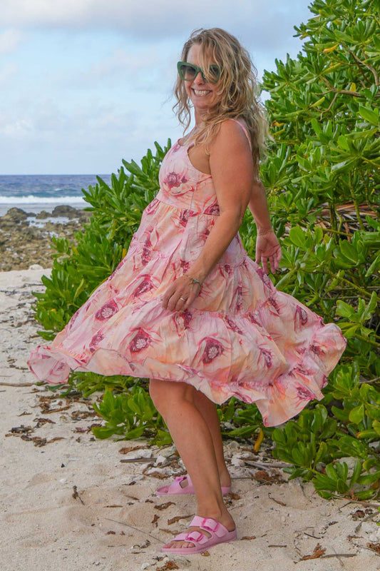 Woman in a pink floral dress standing on a beach with greenery and ocean in the background