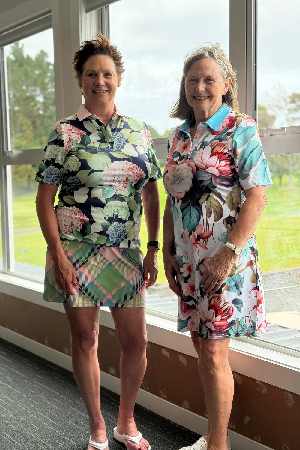 Two women in floral golf dresses standing in a room with large windows.