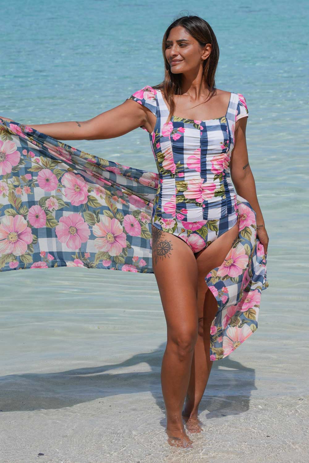 Georgie Bather Gingham Bloom Annah Stretton – front view showing full ruched torso, square neckline, frill cap sleeves, and classic brief cut in Gingham Bloom print, beach setting