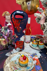 Decorative bottle bag with floral design next to a bottle of wine and festive table setting.